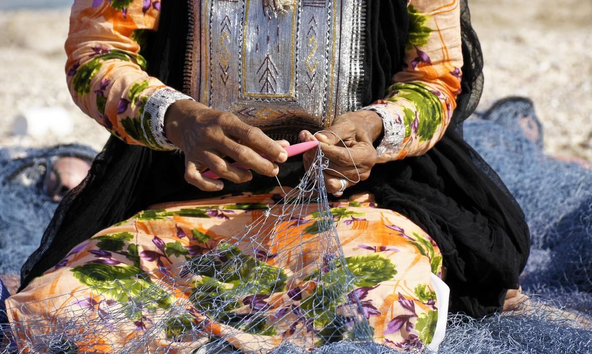 Omani woman in traditional dress mending fishing net on beach, Sur